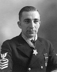 Head and shoulders of a white man with combed-back hair wearing a dark jacket with stripes on the upper sleeve, one round medal pinned to the left breast, and a star-shaped medal hanging from a ribbon around his neck.