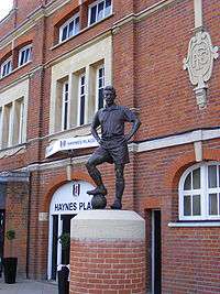 A metal structure composed of a man made of bronze with hands on his hips. One foot is balanced on top of a bronze football. The statue is on an upright brick structure. The word "Haynes" is visible on a sign behind the statue.