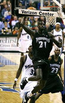 A black basketball player wearing a green jersey prepares to dunk a basketball