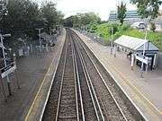 The view back from the footbridge. The pair of 3rd-rail-electrified tracks curve to the left between the 2 platforms. the left one has a sign with the number 2, stairs & a help-point. They both have a shelter, a ticket machine, display boards & lamp-posts. The trees on the left are darker in colour than the trees on the right. There is a silver car on the right & a modern office building.