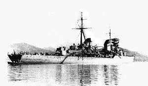 Water-level rear quarter view of a large grey warship at anchor. One gun turret, the main mast and both funnels are prominent.