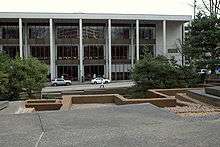 Concrete geometric shapes form a platform in the forefront along with a brown border. In the background are trees and two cars parked in front of a rectangular light-colored building with dark windows.