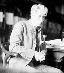 A man in a tweed suit and bowtie sits in a wooden chair beside a desk. He is leaning on the desk and faces to the right but gazes into the camera out of the corner of his eye. His wavy hair is parted, while his hands are folded above his lap.