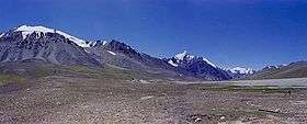 Some tall, bluegrey mountains rise out of brown soil below a deep blue sky