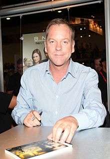 A colored photograph of a blonde-haired man with a blue shirt. He is looking directly at the camera, with a pen on his right hand and book visible on the table.