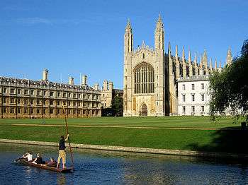 Large yellow stone building with an arched window and two towers at the end nearest the photographer. In the foreground is grass and water with people in a punt.