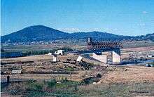 Kings Avenue Bridge under construction. The bridge is incomplete and only the pylons and platform on the southern side are in place. The site is surrounded by dust and the lakebed is dried out.