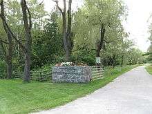 A narrow paved road on the right disappears into the distance. It is lined with trees, and the foreground shows a mown lawn fronting a n open gate, with a wooden fence leading away to the right enclosing the forest behind it.