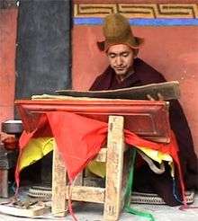 Buddhist monk Geshe Konchog Wangdu in red robe reads Mahayana sutras on stand