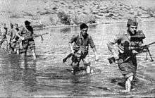 a black and white photograph of a line of males in uniform carrying weapons wading through a shallow river