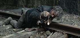 A man with dirty and tousled hair is seen lying on his stomach on a train track, examining a watch that he is holding with his right hand. His shirt is dirty with mud on it and his gloves are torn, baring his fingers. His shoes are also muddy.