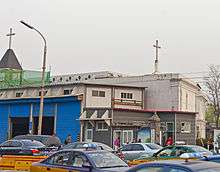 A white two-story building with an irregularly shaped roof seen from a hsort distance away. Two crosses protrude from the roof. There are some other buildings, a sidewalk, and taxicabs in the street in the foreground