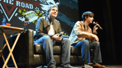 Two men holding microphones sit on a leather couch in front of a large screen.