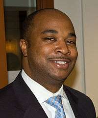 An African-American man wearing a dark suit, white shirt and blue, striped tie