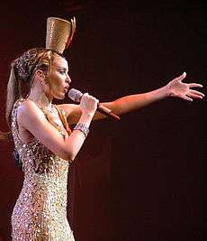 A woman in gold holding a microphone with one arm extended in the air.