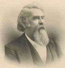 A man with white hair and a white beard and mustache, facing right. He is wearing a white shirt, black vest, and black jacket