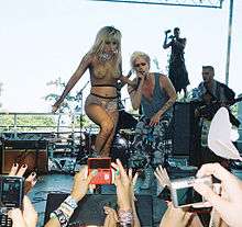 A blond woman stands on a scene while a man, who also has blond hair, is signing behind her..