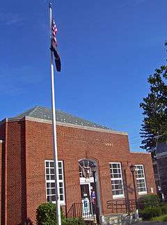 A small brick building with hipped roof and a flagpole in front seen from its left