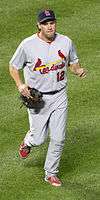 A man in a grey baseball uniform and a dark baseball cap jogs on grass holding a baseball glove in his right hand.