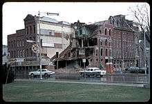 Two roads cross at an intersection in an urban area. At far right, a sign assembly reading west 319 is posted adjacent to one of the roads. In the background, a large building has been partially torn down, resulting in the exposure of the building's interior.