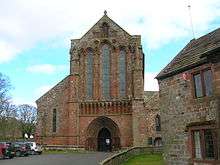 The west end of a red stone church with an elaborate doorway, blind arcading, three tall lancet windows, and a niche containing a statue in the gable