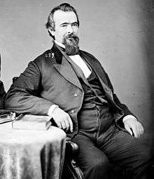 A heavy-set, white-haired man with a beard and mustache, facing right. He is wearing a white shirt and black jacket