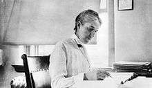 Woman sitting at desk writing, with short hair, long-sleeved white blouse and vest