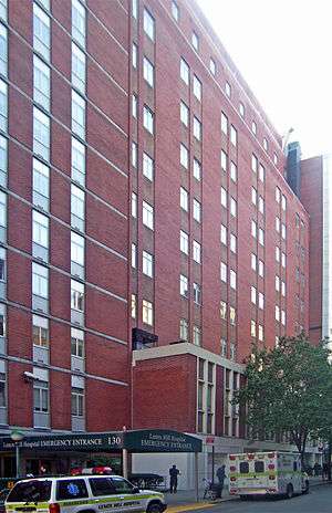 A tall brick building with a dark green awning at street level reading "Lenox Hill Hospital Emergency Entrance". On the street in front are two ambulances and a sport-utility vehicle with flashers in the same color scheme with "Paramedics" on the side.