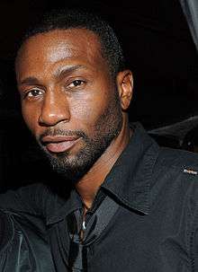 Face of an African-American male with short cropped beard and mustache. He is wearing a black shirt and shiny, black tie.