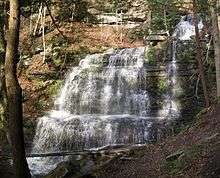 A waterfall spills down a sunlit stone wall made of many layers of rock, surrounded by foliage.