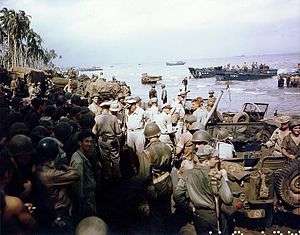 A large crowd of soldiers and jeeps on a beach. There are palm trees in the distance and landing craft offshore. A small group in the center conspicuously wear khaki uniforms and peaked caps instead of jungle green uniforms and helmets.