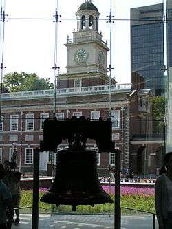 The Liberty Bell hangs in a glass-backed structure, with a brick, 18th-century building with a steeple visible in the background.