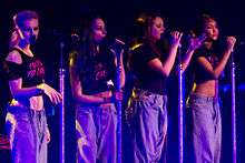 Four women on a stage singing into standing microphones. All women are wearing black tops and grey baggy pants. The women on the far left and far right's tops reveal their stomach areas.