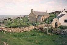 A small windowless church in a churchyard, with the sea visible in the distance. A stone arch is visible over the path leading into the churchyard.