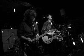 Two guys and a girl playing guitar and another guy playing the drums