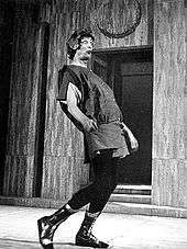 A tall figure in a tunic, black trousers and boots, wearing a laurel wreath and a pop-eyed expression, is moving from left to right in an exaggerated pose, head back and midriff thrust forward. In the background are steps leading to a door.