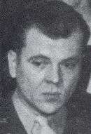 Clean-shaven Caucasian man wearing a suit and tie. He is glancing to the side and has his wavy hair combed back.