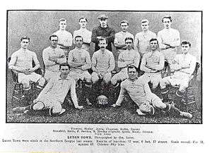 An association football team. Six men stand across the back, the third from the left of which is wearing a dark shirt and a cap. In front of them sits a row of seven men on chairs, with a mustachioed gentleman with hair that is quite long for the time sitting in the middle. Finally, two men recline on the floor in front of them, one on either side of a football marked "LTFC 1909–10".