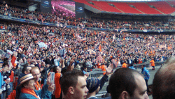 A three-tiered football stadium stand, the bottom two full of people clad mostly in white and orange. Several white and orange flags are visible.