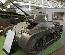 A squat dark green tank in a museum, placed on a large green felt mat. There is a description board in front of it, and a grey divided wall to its rear. The tank's gun barrel is pointing towards the ceiling and its left-hand caterpillar tracks are visible, as well as several large handles on its hull.