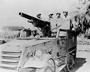 A photograph of a M3 Gun Motor Carriage in a flat area. A mountain is visible in the background, but is obscured by clouds.