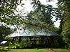 A conical green metal roof supported by many black pillars, the whole structure is in the midst of woods