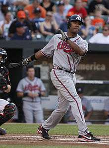 A man, wearing a dark blue batting helmet, a grey baseball uniform with the Atlanta Braves logo across his chest partially obscured, follows through after swinging at a pitch with his bat clutched by his right hand.