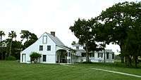 A color photo of a white two-story building with black shutters, a tall wood-shingled roof, and an outdoor veranda.