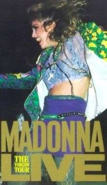 Madonna dancing while looking towards her right. She is clad in a brightly colored jacket and a purple skirt, with a crucifix hanging from her neck.