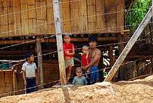 A man and four children stand in front of a wooden hut and behind a wire fence.