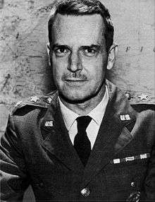 Man with dark hair and moustache in a dress uniform, suit and tie, sitting at a table, with a star indicating his rank, in front of a map on a wall.