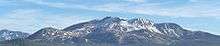 Tall mountain with a wide base and snow interrupted with gray to brown outcrops of rock. Forested area in the fore- to mid-ground.