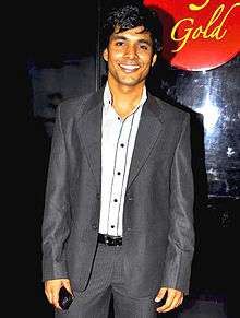 An Indian man smiling, wearing a dark grey suit, with a white shirt under and holding a mobile in his right hand.