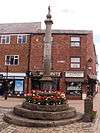 A stone cross on a stepped plinth. It sits on paved ground, with red brick shops behind it.